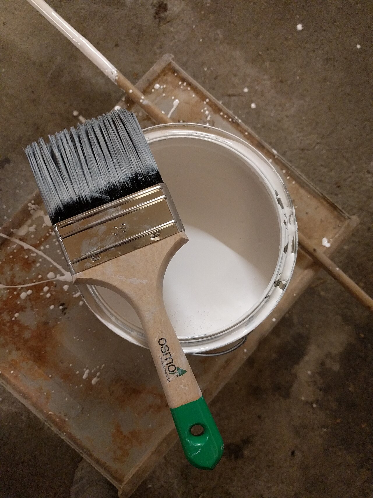A wide paint brush resting on an open can of white paint
